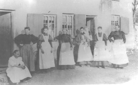 faded photograph of seven ladies in aprons outside a house (source unknown).